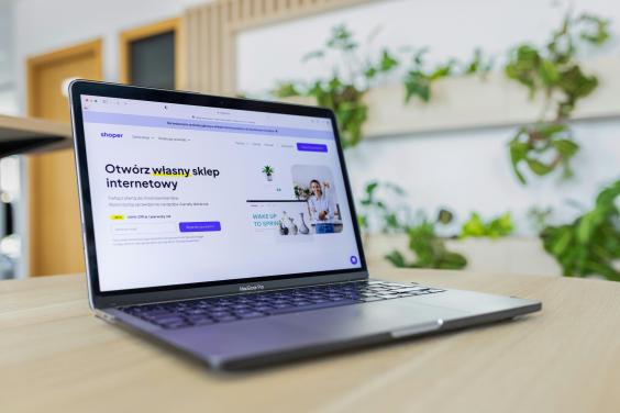 Laptop con p&aacute;gina web de comercio electr&oacute;nico abierta, en un escritorio de madera, con plantas verdes al fondo.